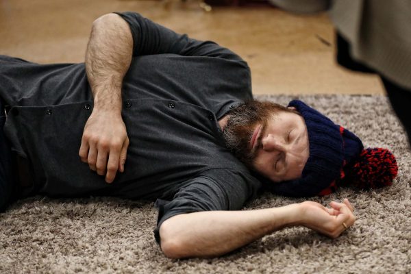 A man with a beard wearing a navy knitted hat and dark shirt is lying on his side on a carpeted floor with his eyes closed, appearing to be asleep or unconscious.