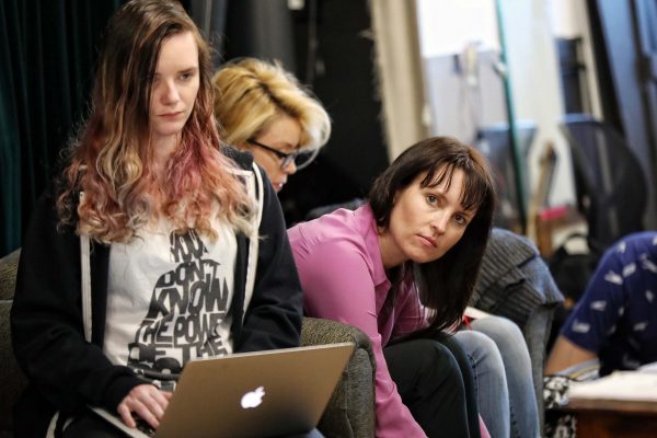 Three women sit indoors; one at the front uses a laptop, another in glasses looks down, and a third in a pink shirt leans forward, gazing attentively to the side.