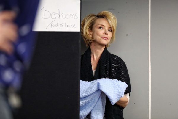 A woman with blonde hair stands next to a sign that says Bedrooms Rest of house, holding a light blue textured blanket and looking to the side with a thoughtful expression.