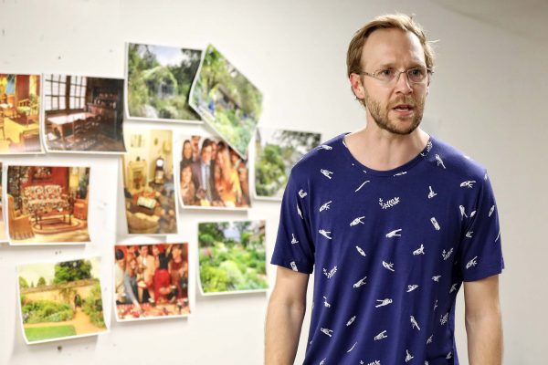 A man with glasses and a blue patterned shirt stands in front of a wall with several colourful photos of gardens, rooms, and groups of people pinned on it. He appears to be talking or presenting.