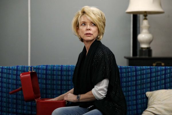 A woman with short blonde hair sits on a blue checked sofa, looking to the side while holding an open red box on her lap. There is a table with a lamp and a cushion in the background.