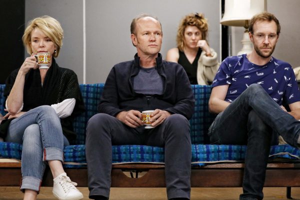 Three people sit on a blue checked sofa holding mugs, looking serious. A fourth person sits in the background, also looking thoughtful. The scene suggests a tense or contemplative moment.