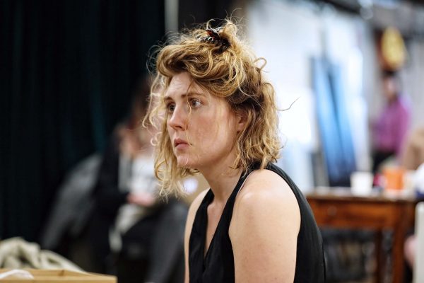 A woman with wavy, tousled blonde hair and a pensive expression sits indoors, wearing a sleeveless black top. The background is blurred, showing indistinct people and furniture.