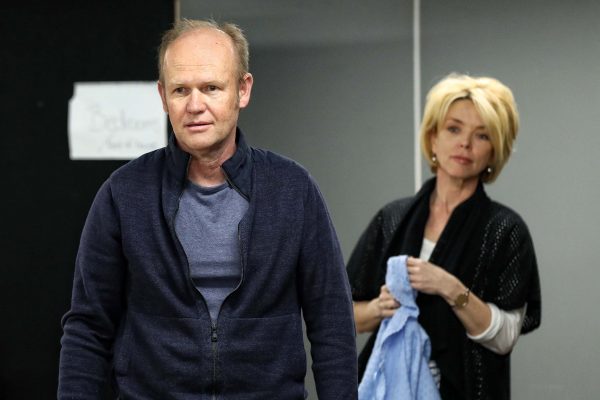 A man in a blue jacket stands in the foreground, while a woman holding a blue shirt stands behind him. Both have serious expressions and are indoors with a blurred background.