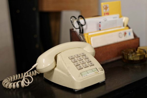 A beige rotary-style landline telephone sits on a dark surface next to a wooden organiser holding letters, envelopes, and a pair of scissors. An ashtray is also visible on the right side of the image.