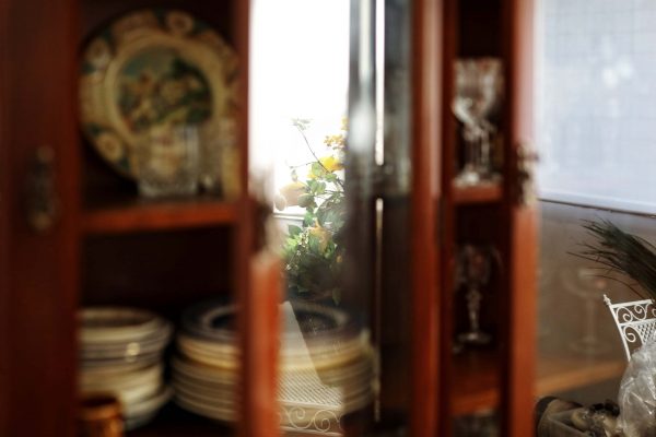A glass-fronted wooden cupboard displays stacked plates and decorative glassware. Through the glass, yellow flowers and sunlight are visible in the background, casting a warm, cosy ambience.