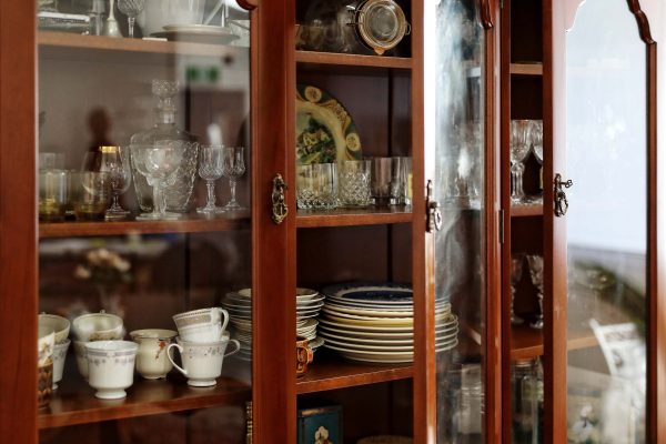 A wooden china cabinet with glass doors displays assorted glassware, teacups, teapots, and stacked plates on its shelves. The arrangement is neat, with reflective surfaces enhancing the elegant, vintage look.