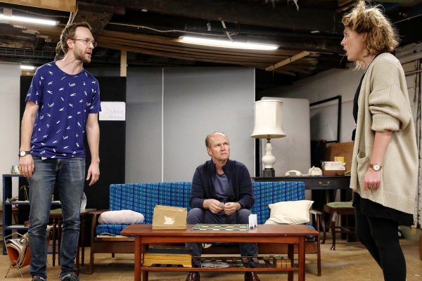 Three people are in a cluttered room. Two are standing and appear to be arguing, while a third person sits on a blue sofa between them, looking concerned. There is a coffee table with items on it in front of the sofa.