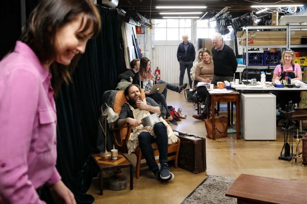 A group of people in a casual, creative workspace. Some are sitting and chatting, one woman is laughing on an armchair, and another stands smiling in the foreground. Various work materials and computers are visible in the background.