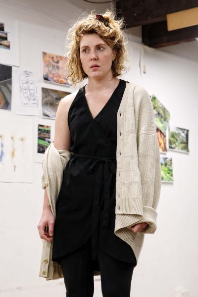 A woman with curly blonde hair wears a black dress, black leggings, and a loose beige cardigan. She stands indoors against a white wall with various drawings and photos displayed behind her.