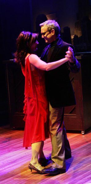 A woman in a red dress and a man in a dark jacket and glasses dance together on a dimly lit stage, holding hands and gazing at each other.