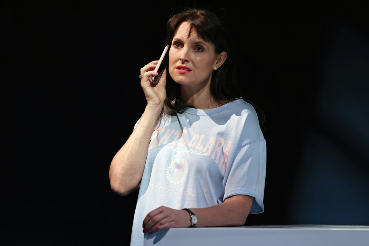 A woman with long brown hair in a ponytail, wearing a light blue T-shirt, holds a phone to her ear and looks thoughtful, standing against a dark background.