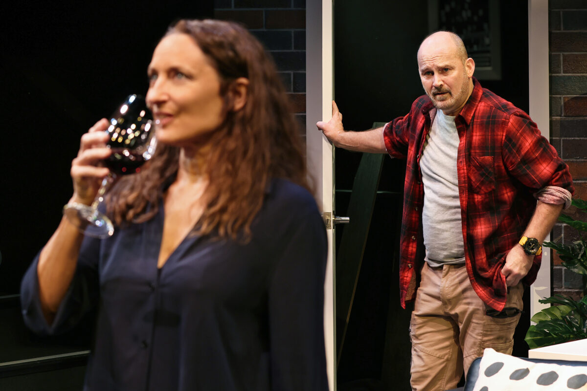 A man in a red checked shirt stands by an open door, looking concerned, while a woman in the foreground, out of focus, sips from a wine glass. They appear to be on a stage set.