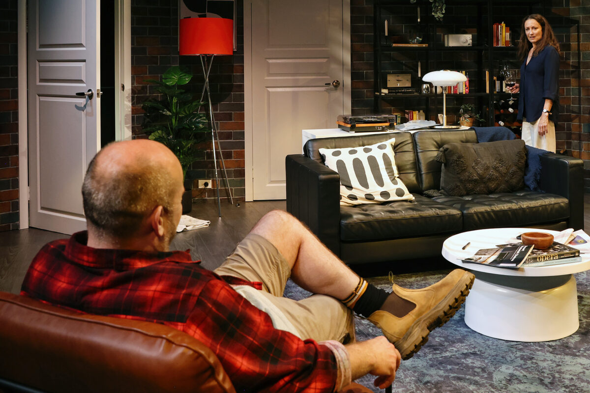 A man in a red checked shirt sits on a chair facing a woman standing by a bookcase in a modern living room with a black sofa, round white table, and decorative items.