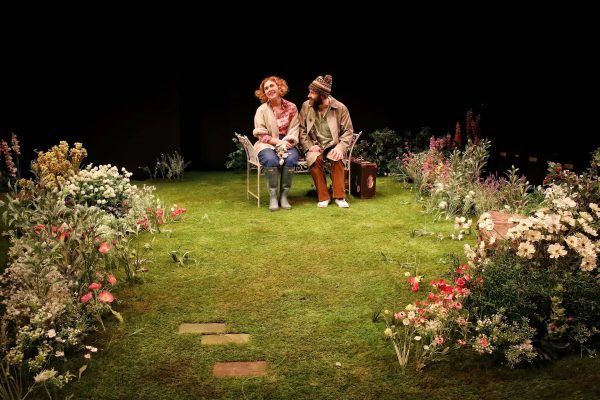 Two people sit on a bench in a lush, flower-filled garden under stage lighting. The scene is vibrant with various colourful flowers and greenery, and the pair appear to be engaged in a conversation.