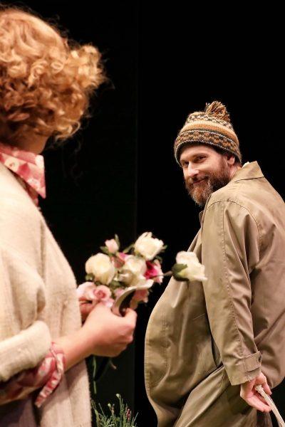 A man in a beanie and beige coat smiles playfully at a woman holding a bouquet of white and pink flowers. The background is dark, highlighting the two figures.