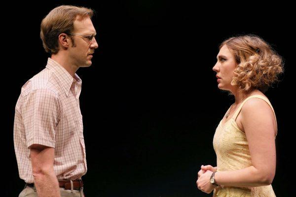 A man in glasses and a checked shirt faces a woman in a light yellow dress. They stand close together, looking at each other with serious expressions against a plain, dark background.