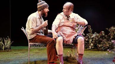 Two men sit on a garden bench surrounded by plants; one wears a knitted hat and holds a bottle, the other, in a pink apron and dress shoes, holds a glass and newspaper. They appear to be having an animated conversation.