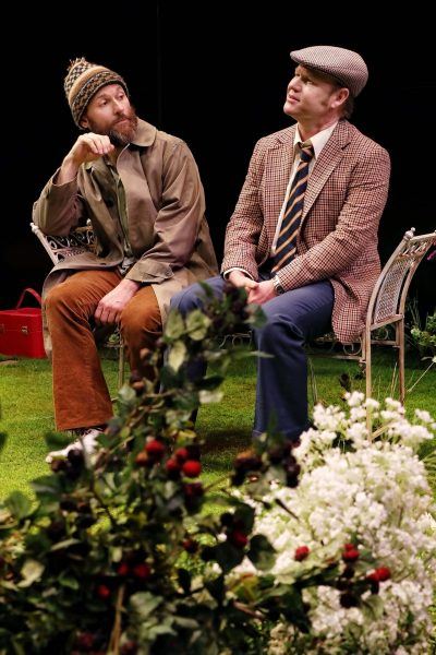 Two men sit on metal chairs in a garden setting. One wears a beanie, brown jacket, and corduroy trousers; the other wears a flat cap, checked blazer, blue trousers, and tie. Flowers and greenery fill the foreground.