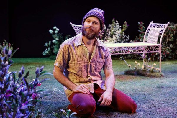 A bearded man in a knitted hat and short-sleeved shirt kneels on grass in a garden setting, surrounded by flowers, with a white metal bench and foliage in the background, under stage lighting.
