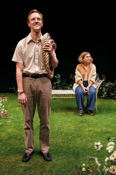 A man in glasses holds a coiled rope and stands on a grassy stage, smiling. Behind him, a woman sits on a white bench, looking off to the side. Flowers and greenery are scattered across the stage.