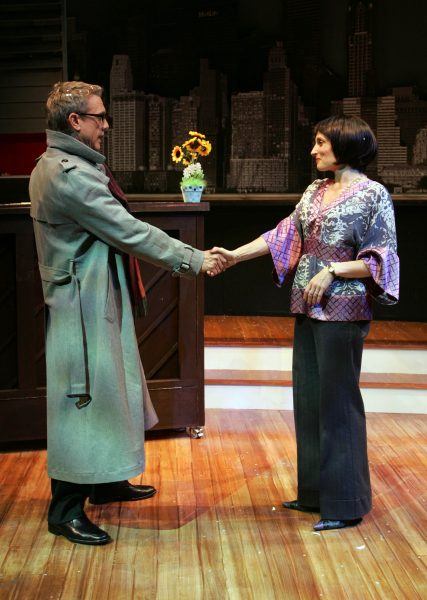A man in a grey trench coat and sunglasses shakes hands with a woman in a patterned blouse on a wooden stage, with a cityscape backdrop and a potted plant on a counter behind them.