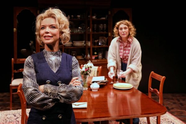 Two women are in a dining room; one stands confidently in the foreground with arms folded and a smile, while the other stands behind a wooden table, arranging crockery and looking forwards.