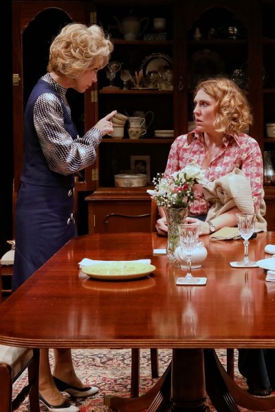 Two women in vintage clothing are in a dining room. One stands, pointing sternly, whilst the other sits at the table looking upset, clutching a serviette. The table is laid with flowers, glassware, and empty plates.