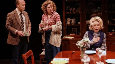 Three actors perform a dramatic scene in a dining room; a man in a checked jacket and tie stands holding a dish, a woman in a floral blouse looks concerned, and an older woman sits at the table with her hand on her chest.
