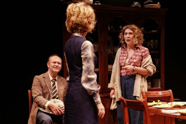 Three actors perform a dramatic scene in a dining room setting. A man sits holding a cabbage, while two women stand; one looks surprised, the other faces away. A table with food and a wooden cupboard are in the background.