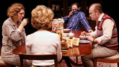 Four adults sit around a wooden dining table set with breakfast items, including toast, tea, and cereal. One man covers his nose with his shirt, while the others look serious or contemplative.