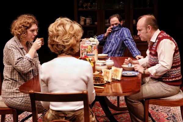 Four adults sit around a wooden dining table set with breakfast items, including toast, tea, and cereal. One man covers his nose with his shirt, while the others look serious or contemplative.