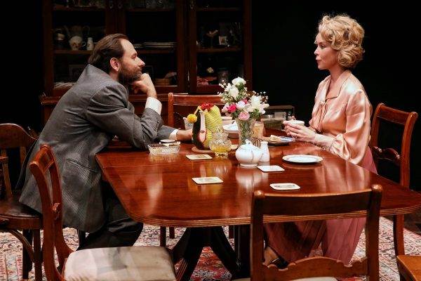 A man in a suit and a woman in a pink dressing gown sit opposite each other at a wooden dining table set with flowers, cups, and dishes, appearing to have a serious conversation in a warmly lit room.