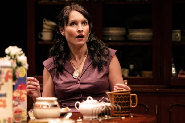 A woman with long dark hair, wearing a purple sleeveless blouse and a pendant necklace, sits at a table set with tea things, looking surprised or shocked, with crockery and a dark cupboard in the background.