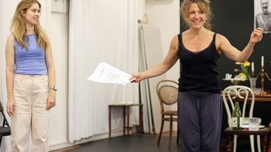 Two women stand in a rehearsal room. One, holding a script and gesturing, smiles enthusiastically. The other, dressed in light trousers and a blue top, looks on. The background shows chairs, a table, and a blackboard.