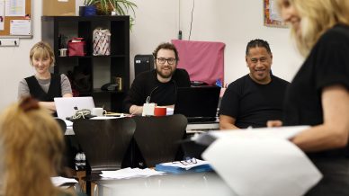 Three people sit at a table with laptops and papers, smiling and looking towards a woman standing in the foreground who is holding documents. The setting appears to be a casual office or classroom environment.