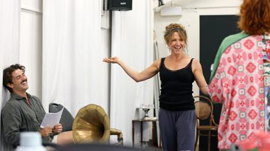 A woman stands smiling with arms outstretched, engaging with a person in colourful clothes. A man sitting nearby laughs, holding a script. The setting appears to be a casual rehearsal studio.