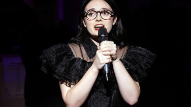A young woman with dark hair and glasses, wearing a frilled black dress, holds a microphone with both hands and sings under dramatic stage lighting.
