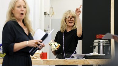 Two women are indoors; one holds papers and looks surprised, while the other, wearing glasses and holding a wrapped box, stands behind a wooden table, pointing upwards and speaking animatedly. Papers and a red mug are on the table.
