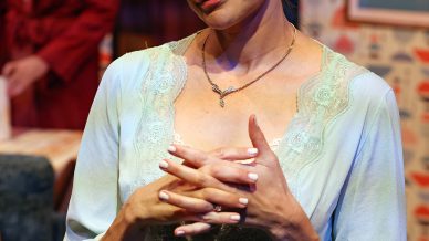 A woman in a light blue dressing gown stands with her hands clasped, smiling softly. She is indoors with patterned wallpaper and framed art. A man in a red dressing gown is blurred in the background.