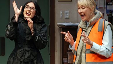 Two women stand indoors. One wears a black dress and glasses, gesturing expressively. The other, in a reflective orange waistcoat, smiles and points with both hands. They appear to be interacting enthusiastically.
