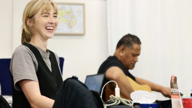 A woman smiles brightly whilst sitting at a table with papers and a laptop; in the background, a man plays an acoustic guitar. The scene appears to be in a casual or creative workspace.
