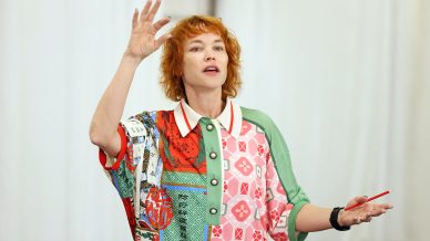 A person with curly red hair wearing a colourful, patterned shirt gestures with one hand raised and holds a red pen in the other, standing in front of a light, blurred background.
