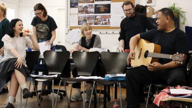 Six people are gathered in a bright room with chairs, papers, and a guitar. They are smiling and talking, with one person playing the guitar. A noticeboard with photos and notes is visible in the background.