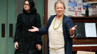 Two women stand indoors; one in a black dress with glasses looks ahead seriously, while the other, wearing a light outfit and a green necklace, gestures and smiles. A piano and noticeboard are in the background.