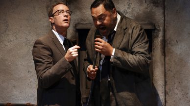 Two men in suits stand close together in front of a rough, grey wall. One man looks up and gestures with a small object, while the other leans forward, shouting into a device with an intense expression.