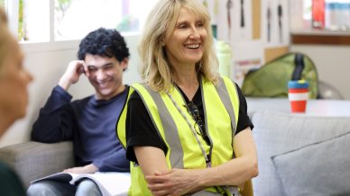 A smiling woman in a yellow safety vest sits on a sofa, with a young man in the background holding a book and smiling. The setting appears to be a casual meeting or workshop in a bright, informal space.