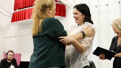 Two women stand facing each other and smiling, holding each other's arms warmly. One wears a green shirt, the other a white blouse. Other people are visible in the background in a bright, indoor setting.