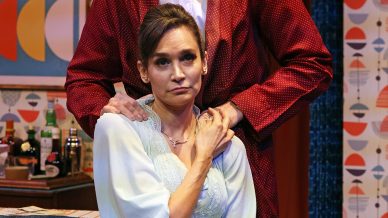 A man in a red dressing gown stands behind a seated woman in a light blue nightdress, gently holding her shoulders. They appear serious, set in a colourful, retro-styled living room with patterned wallpaper and framed art.