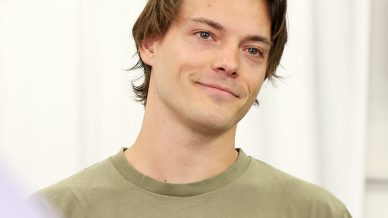 A young man with medium-length brown hair, wearing an olive-green T-shirt that says Thrills Some Kind of Paradise Company, stands indoors, smiling softly. A blurred hand holding a piece of paper is in the foreground.